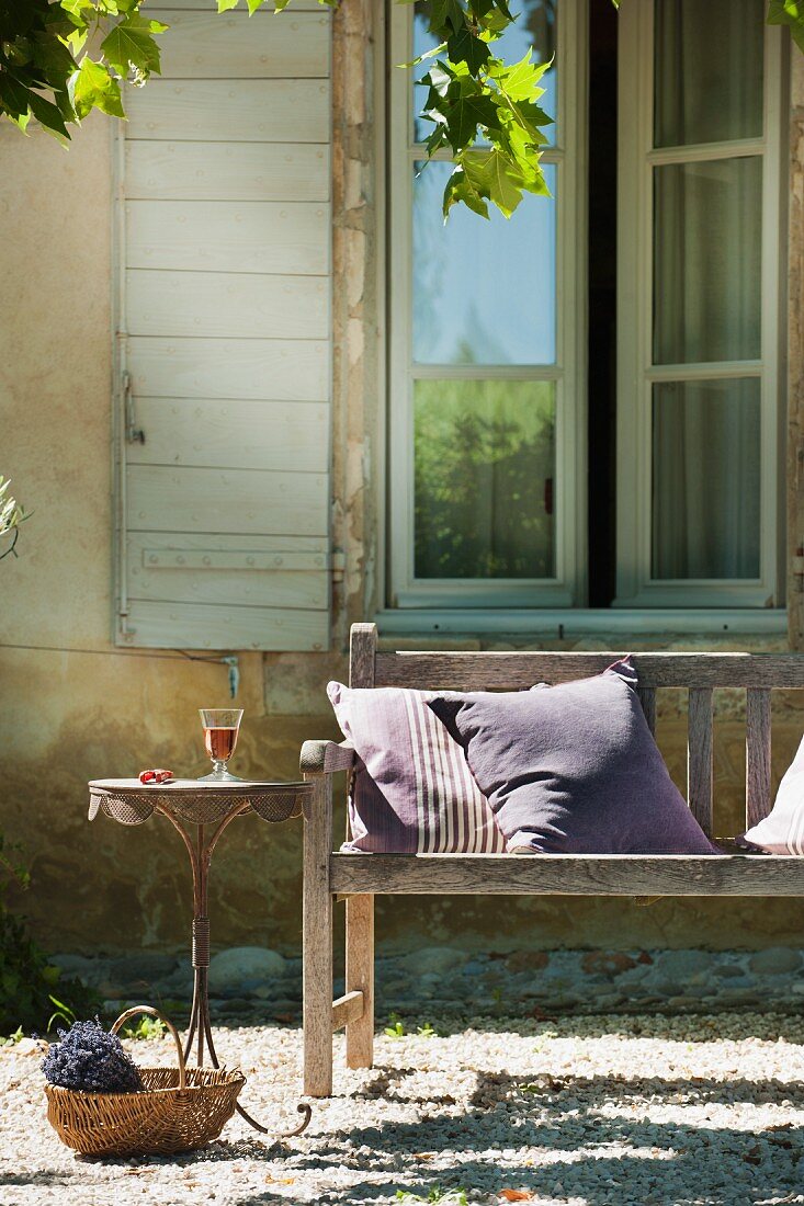 Kissen auf Holzbank, zierlicher Bistrotisch und Korb mit Lavendelstrauss vor dem Fenster eines provenzalischen Landhauses