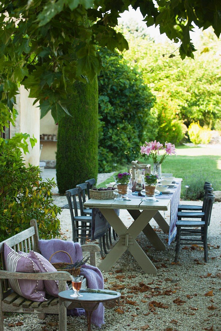 Gedeckter rustikaler Tisch und kleine Bank mit Kissen auf einem Kiesplatz in mediterranem Garten