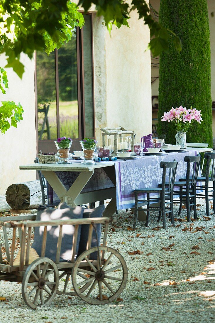Mit blauer Tischdecke und Blumen geschmückte Tafel auf Kiesfläche vor französischem Landhaus; Leiterwagen mit Kissen im Vordergrund