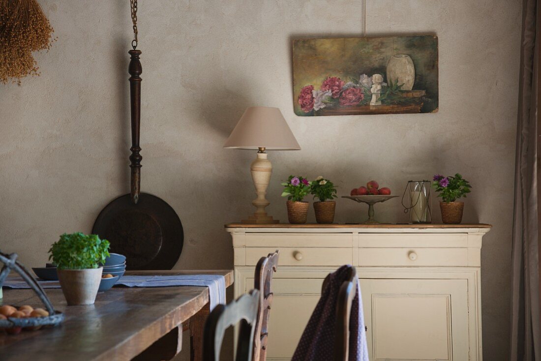 Provenzalisches Esszimmer mit Eisenpfanne und Stillleben an der Wand; schlichte Kommode mit Blumenschmuck und rustikaler Holztisch