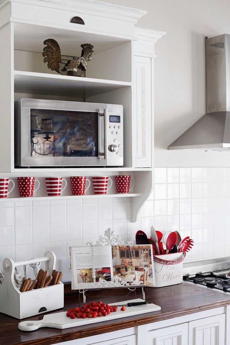 Microwave in a white lacquer floating cabinet on a wall above a kitchen counter with utensils and cook book on a wooden cutting board