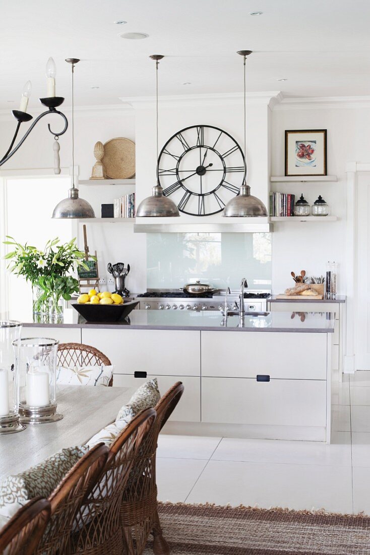 White, open-plan kitchen with pendant lamps, wall clock, rustic dining table and wicker chairs