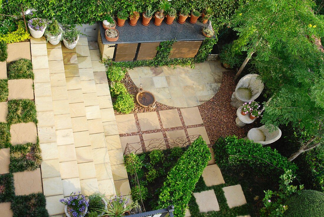 View down onto precise garden with stone-floored seating area, rows of flower pots and beds between stone flags