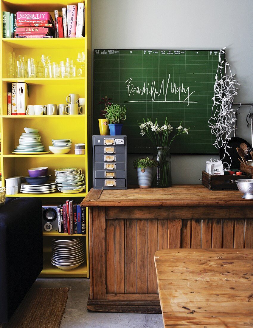 Rustic, half-height cabinet below green chalkboard next to yellow-painted crockery shelves