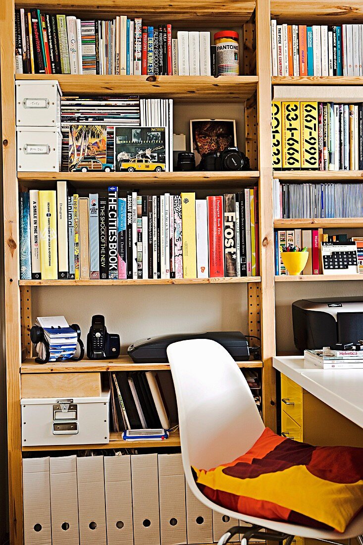 Volle Regalwand in Arbeitszimmer mit weißem Schalenstuhl und weißem Schreibtisch; auf dem Stuhl ein buntes Kissen