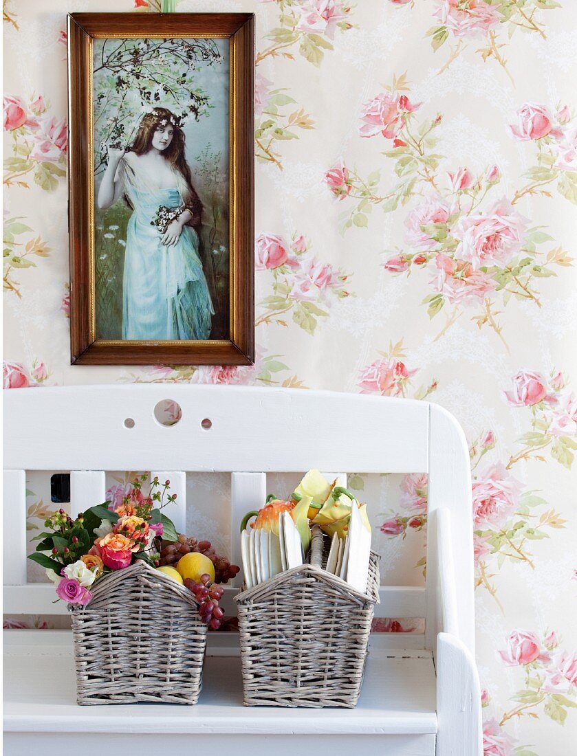 Framed photograph above baskets of garden roses on white wooden bench against rose-patterned wallpaper