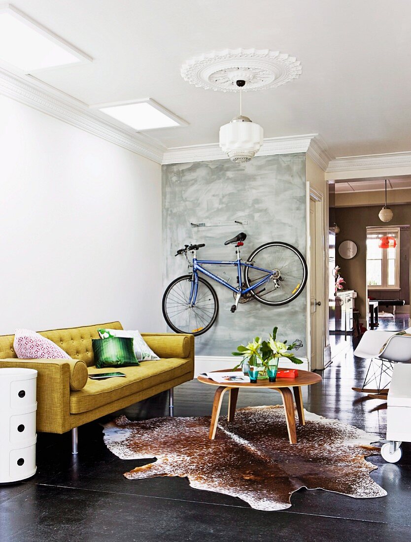 Offener Wohnbereich mit aufgehängtem Fahrrad an der grauen Wand, im Vordergrund Retrosofa mit Tisch auf schwarzem Dielenboden