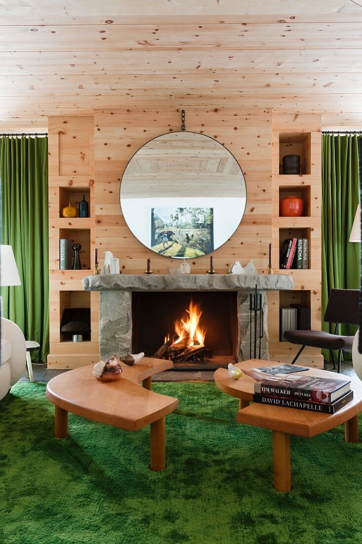 Two-part coffee table on green rug in front of open fire below circular mirror on wooden wall