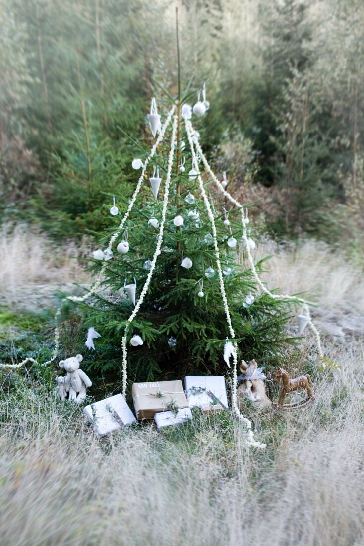 Mit weissen Kugeln, Girlanden und Geschenkpaketen geschmückter Weihnachtsbaum auf einer Waldlichtung