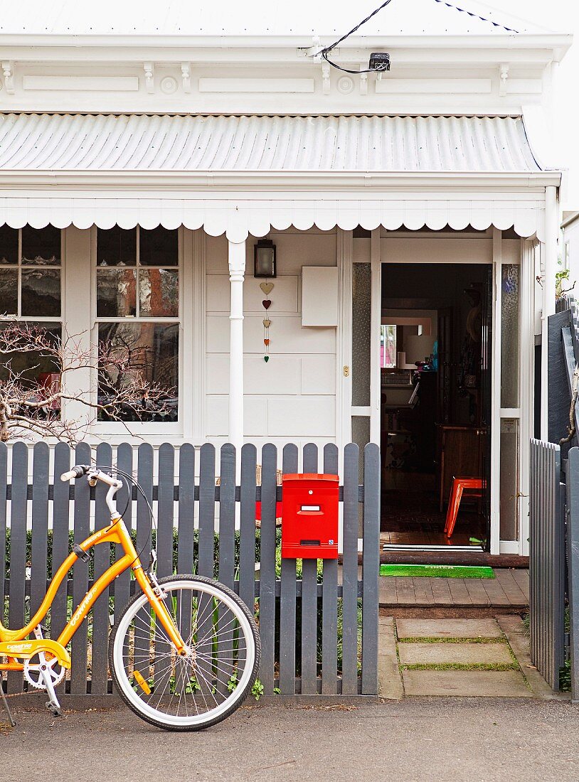 Eingangsbereich mit Vordach zu einem weiß gestrichenen Cottage, am grauen Gartenzaun mit rotem Briefkasten lehnt ein gelbes Fahrrad