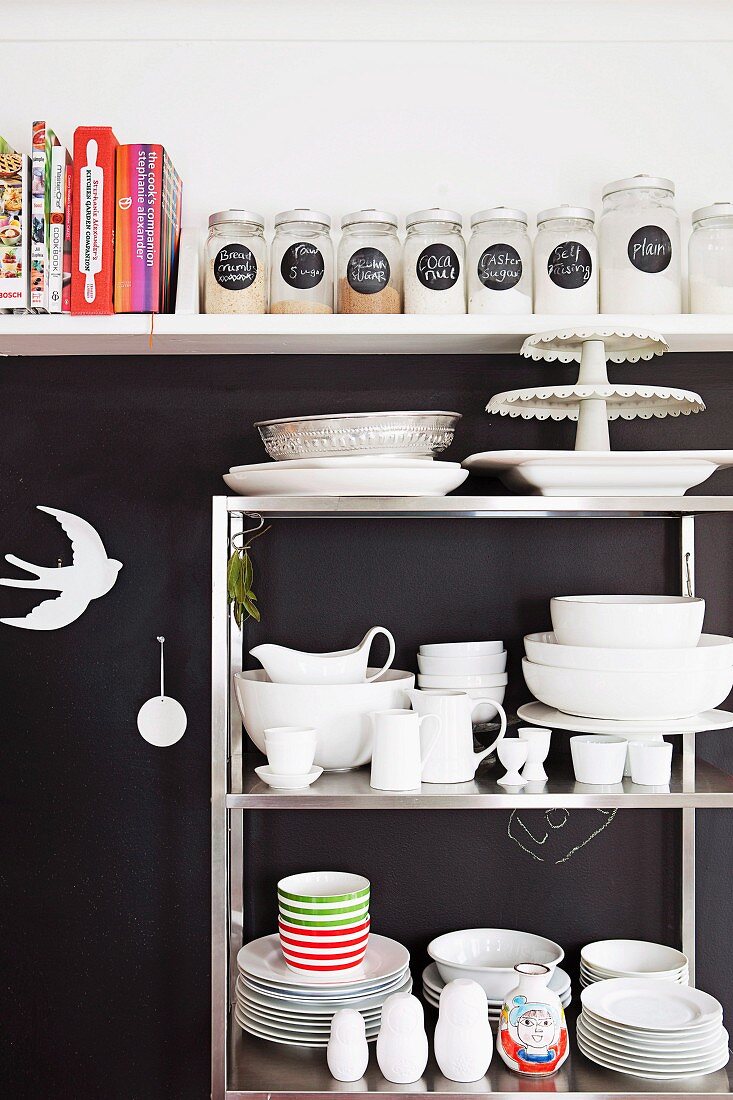 Metallregal mit weißem Geschirr vor schwarzer Wand, darüber weißes Küchenboard mit Kochbüchern und Aufbewahrungsgläsern