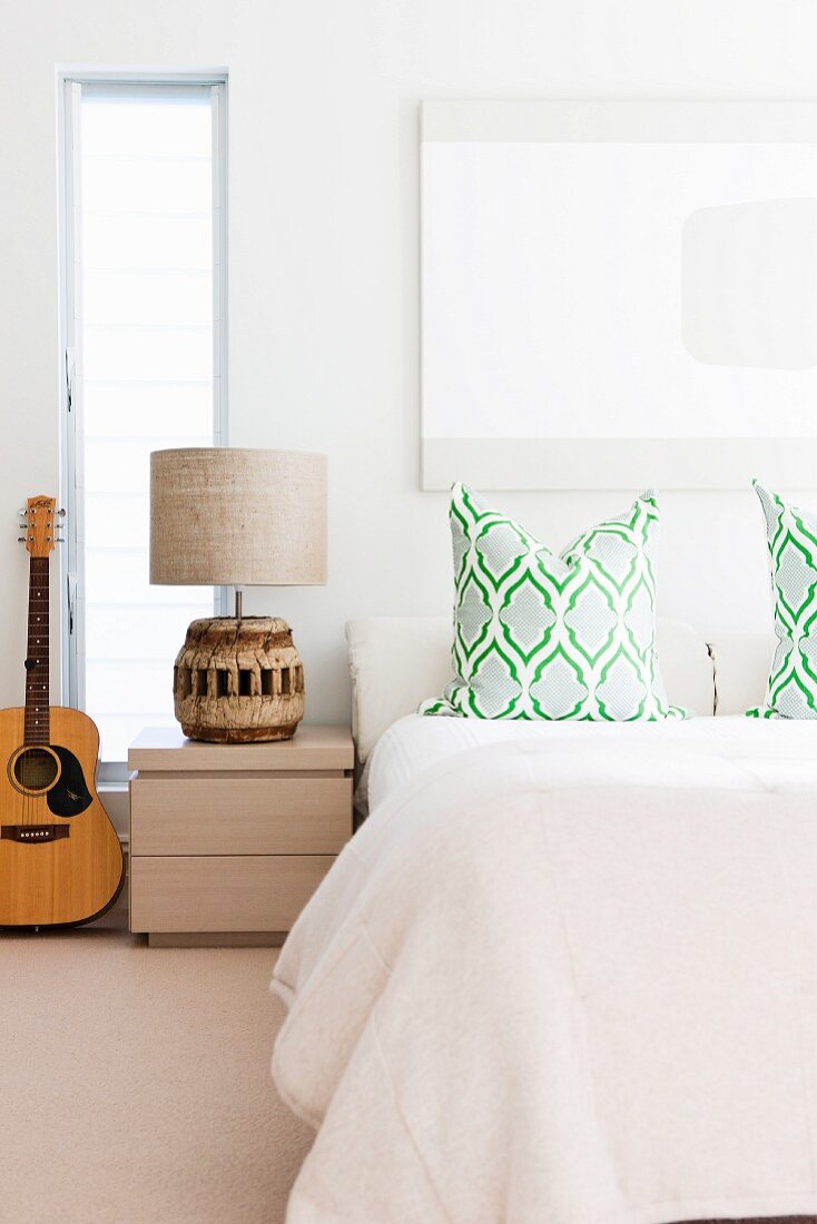 Doppelbett mit Zierkissen; senkrechter Fensterschlitz hinter folkloristischer Nachttischlampe und Gitarre