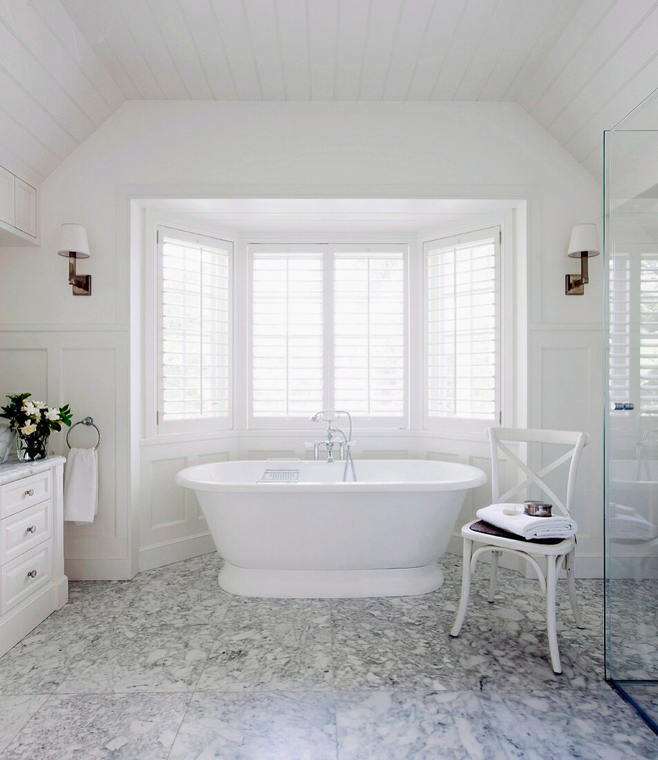 Weißes holzverkleidetes Badezimmer mit Carrara Marmorboden und freistehender weißer Badewanne vor dem Erkerfenster