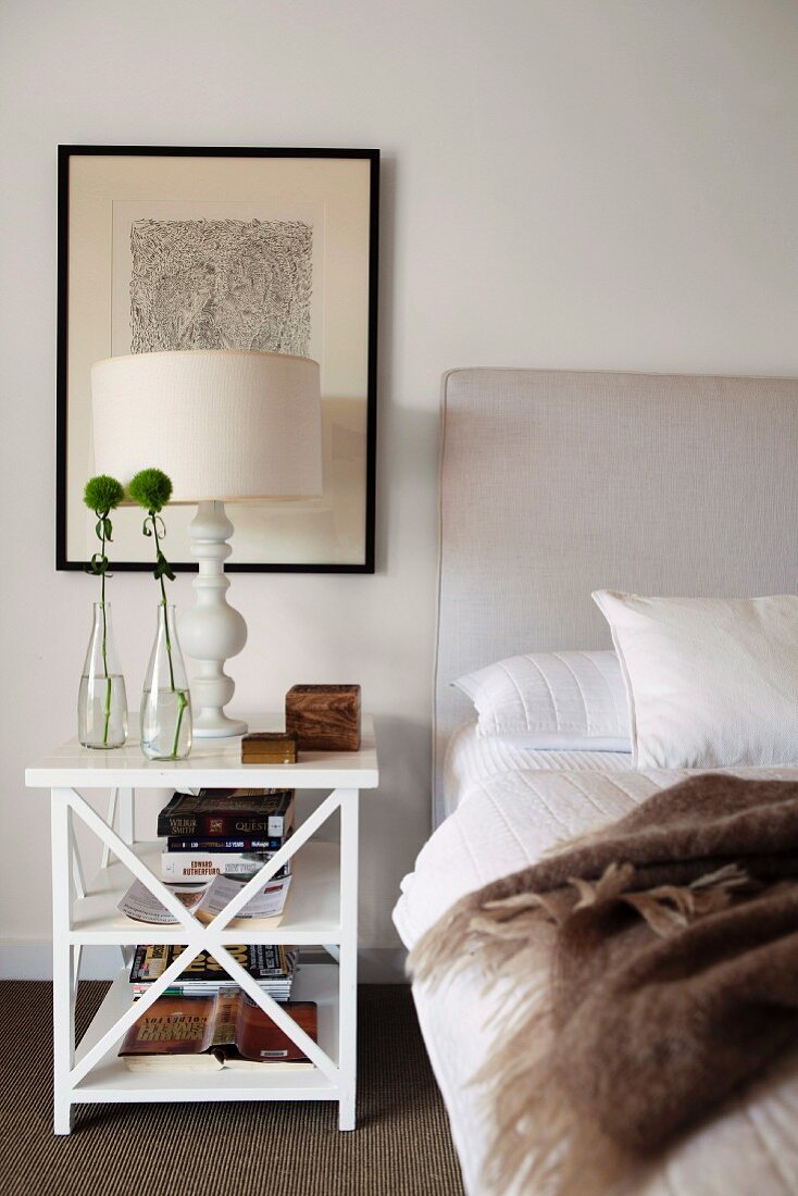 Table lamp on white, wooden bedside table below picture and blanket on bed