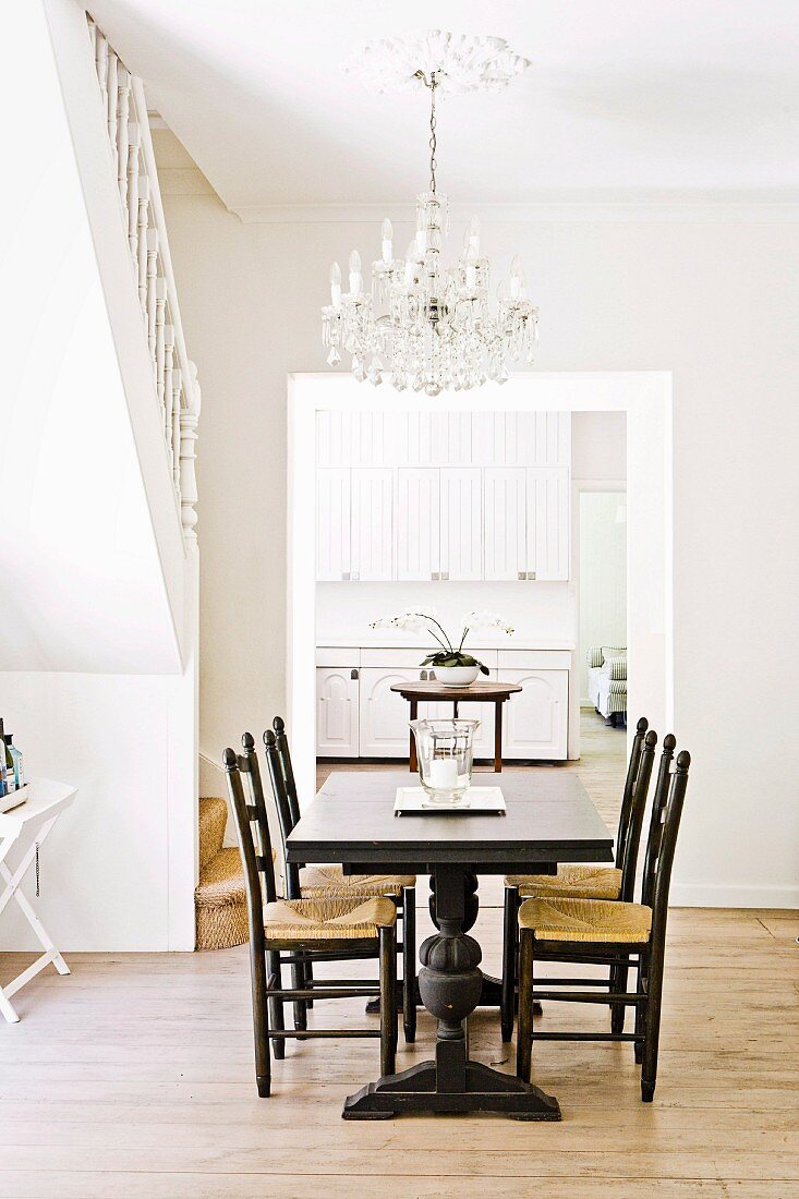 Dark dining table and chairs in bright room with chandelier and open doorway leading to white kitchen