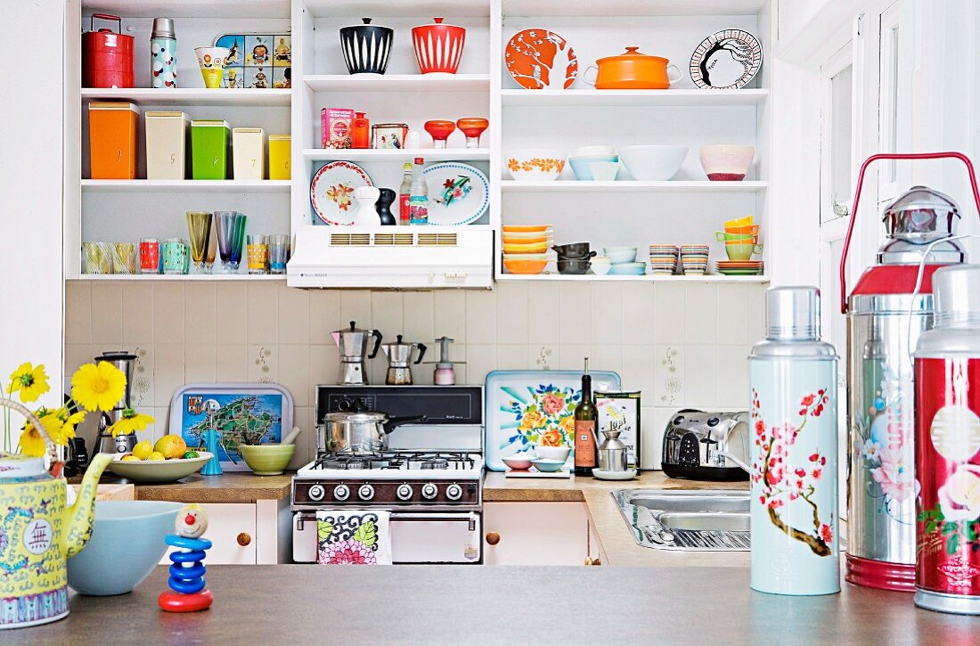 Colourful crockery on white kitchen shelving; brightly coloured thermos flasks on worksurface in foreground