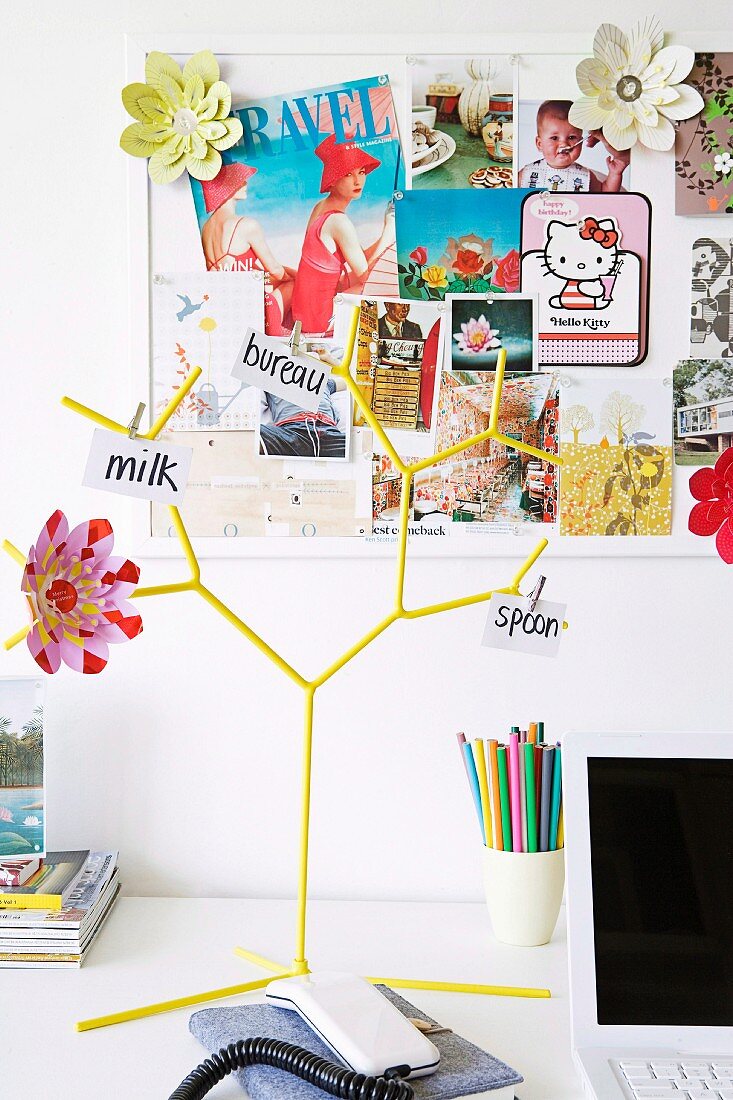 Pinwand mit bunten Postkarten und Dekoblüten, davor ein gelber Tischständer mit Notizen