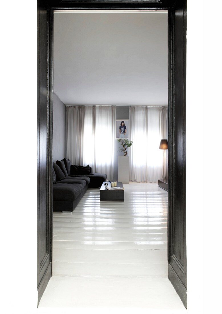 Blick durch schwarze Doppeltür in puristisches Wohnzimmer mit weiss glänzendem Dielenboden und dunkler Sofagarnitur vor verhangenen Fenstern