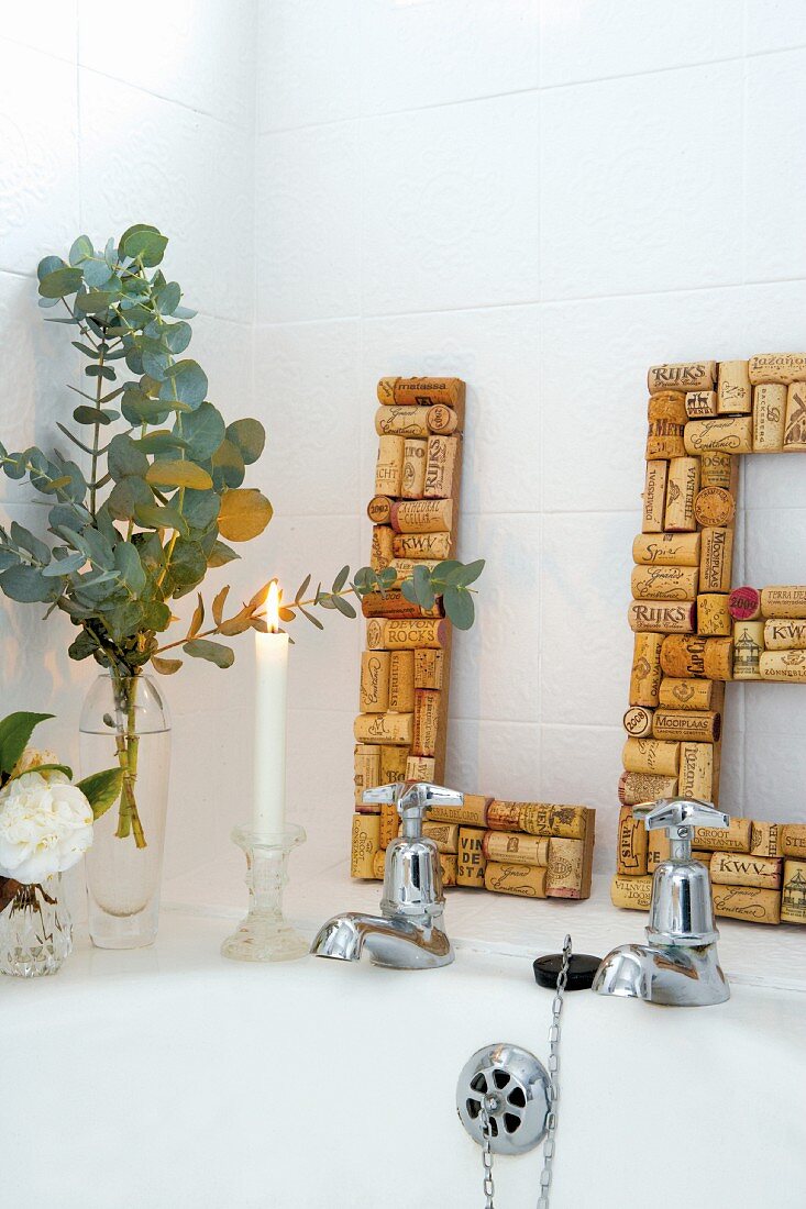 Badewannenrand mit Kerzenlicht und Blumenvasen vor selbstgemachten Korkbuchstaben an weiße Fliesenwand gelehnt
