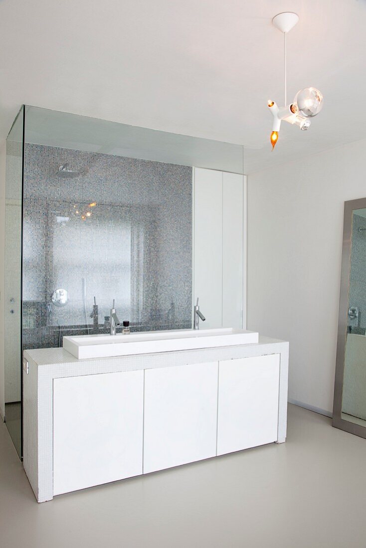 Long masonry washstand with mosaic tiles and long sink against glass partition screening shower area