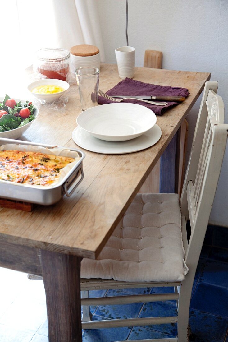 Pikanter Auflauf, Salat und Einzelgedeck aus weißem geschirr auf schlichtem Holztisch mit Küchenstuhl