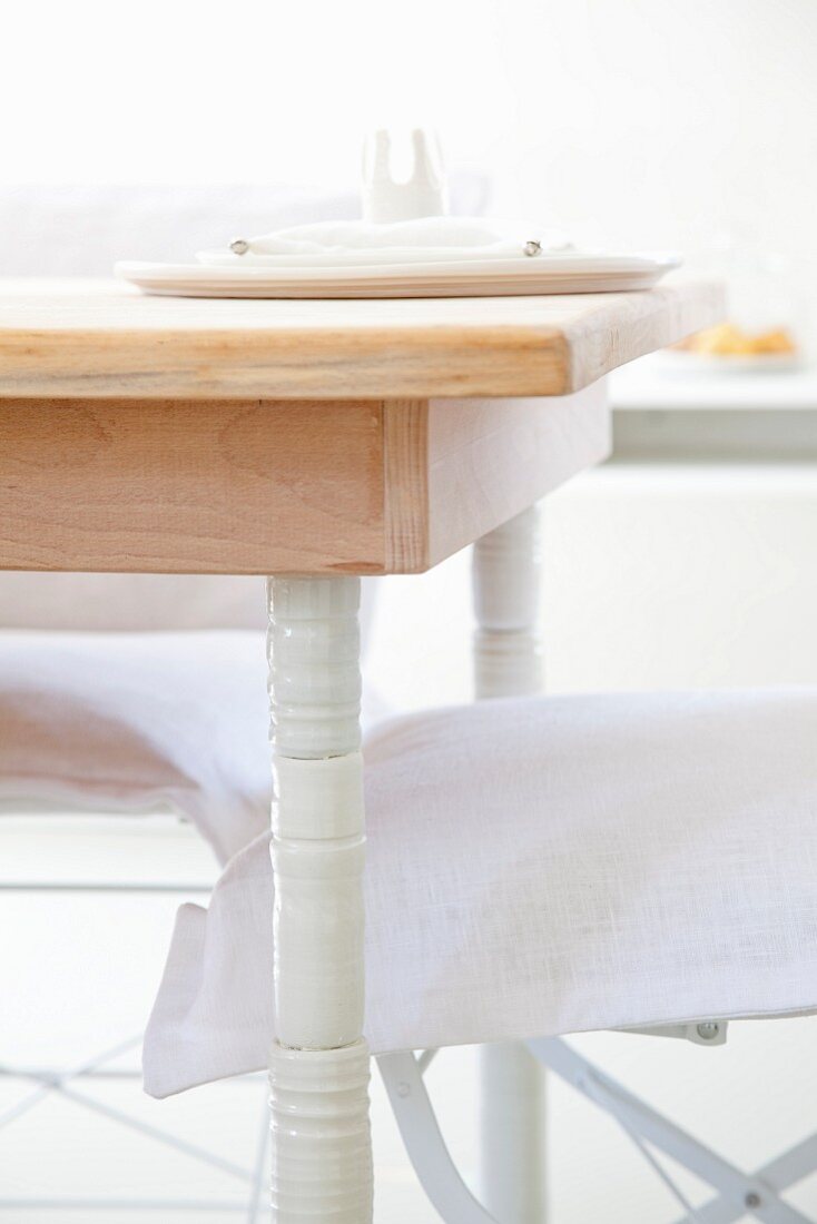 Ecke eines hellen Holztischs mit weiss lackierten Beinen und Gartenstühle mit Sitzkissen