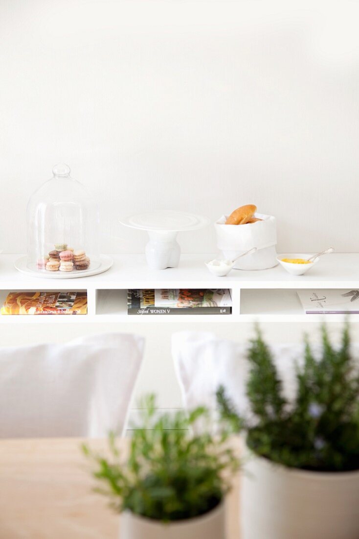 Käseglocke, Kuchenplatte und Tütenschale auf Ablage, Tisch mit Kräutern unscharf im Vordergrund