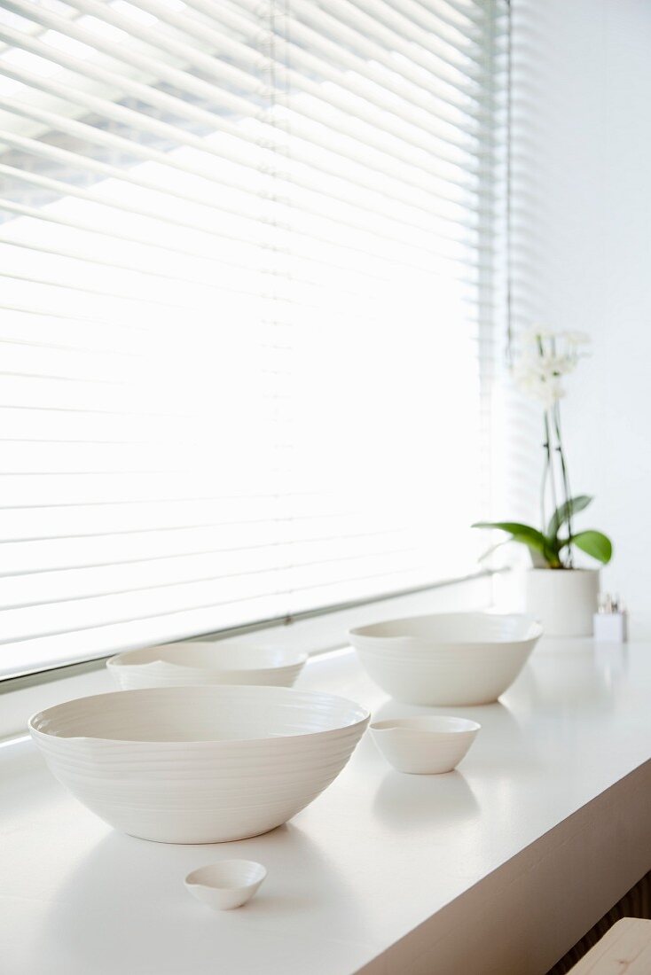 White, hand-made clay dishes of different sizes and potted orchid in soft light falling through half-open louver blind