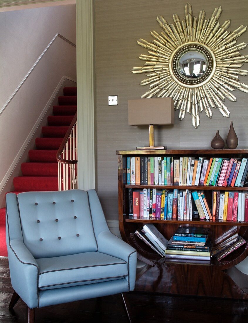 Blue armchair next to half-height bookcase below mirror with sunburst gilt frame on wall