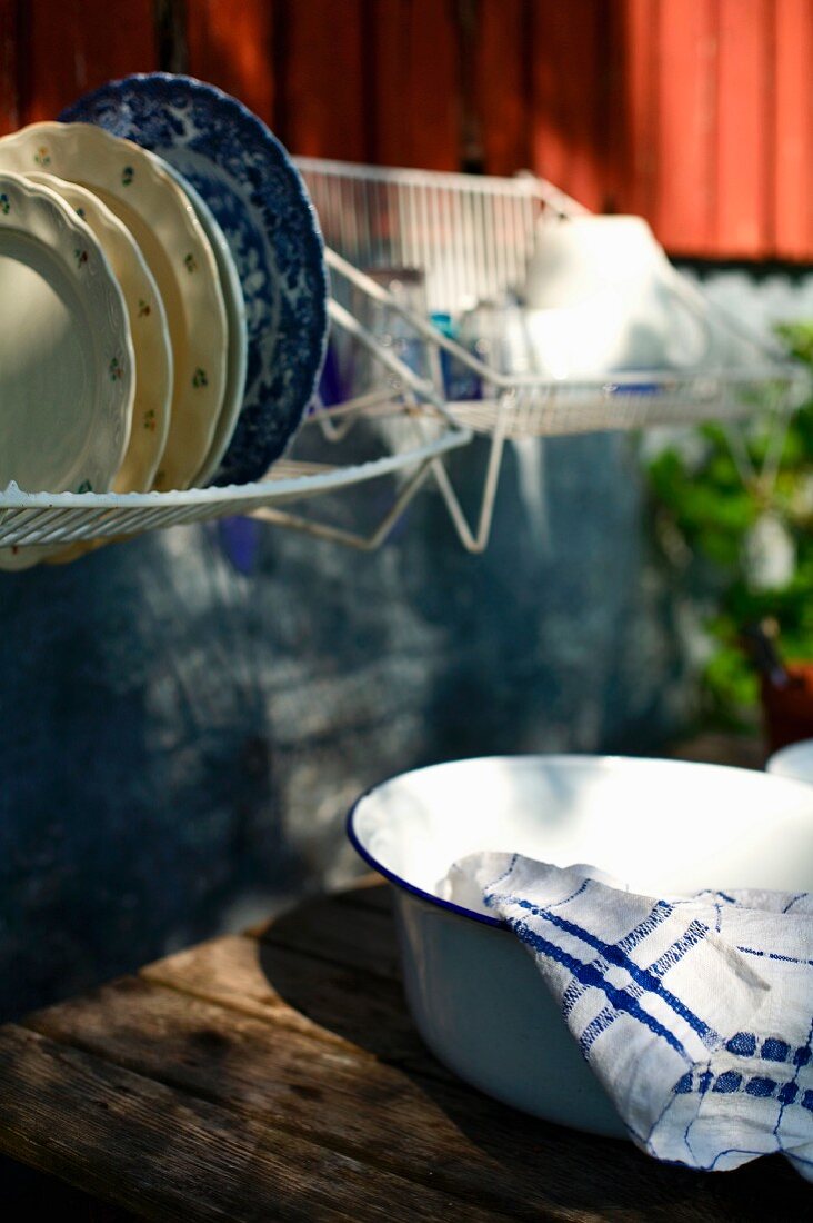 Washing-up bowl on wooden table below drying rack of crockery hung on house facade