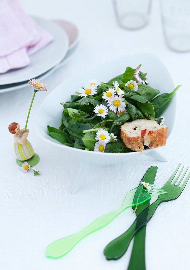 Green plastic cutlery and ornament of girl next to bowl of spinach salad with daisies