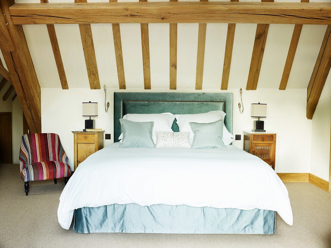 Elegantes Doppelbett mit grünblauer Husse und gepolstertem Kopfteil im Dachzimmer mit Holzbalkendecke