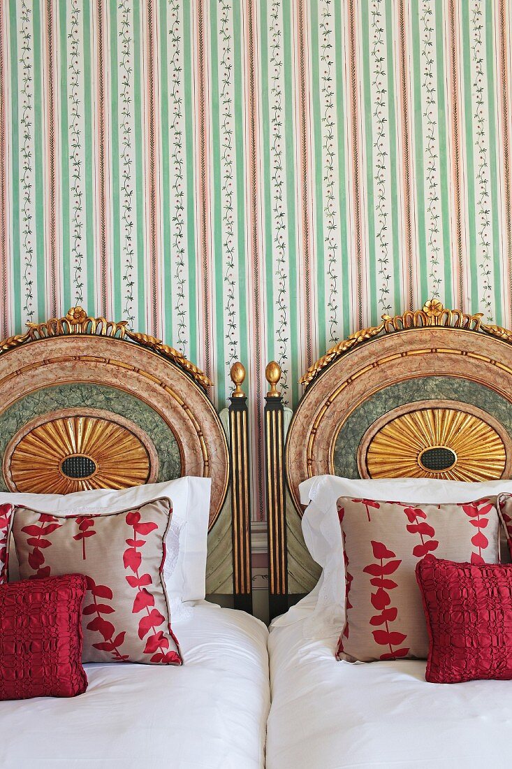 Twin beds with elaborately carved and painted headboards against striped wallpaper