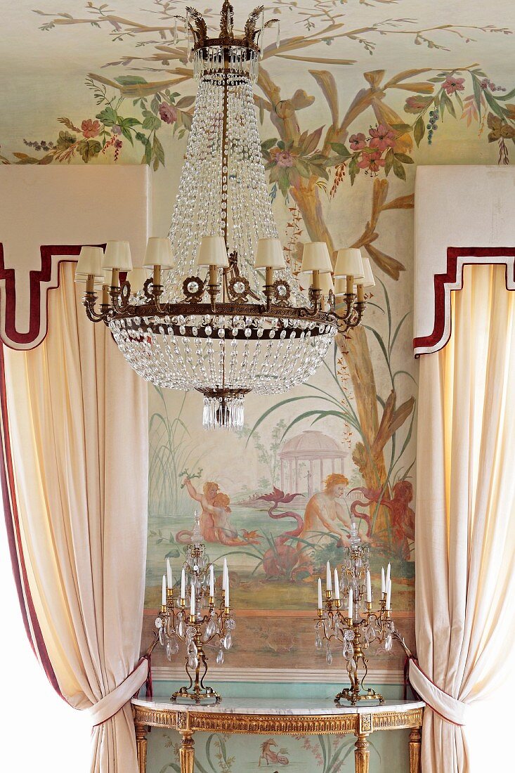 Kronleuchter mit kleinen weissen Schirmen vor bemalter Wand und Kerzenständer auf goldverziertem Konsolentisch