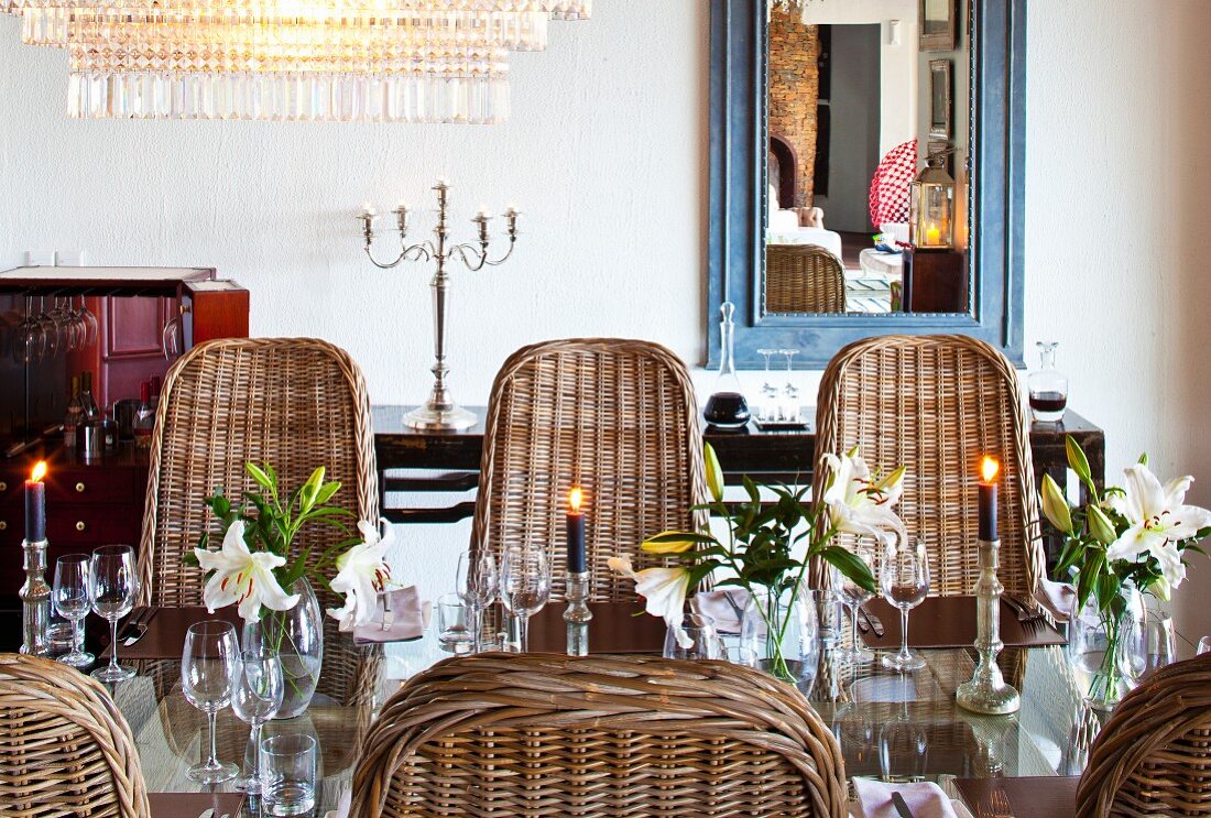 Glänzender Glastisch mit silbernen Kerzenhaltern und Liliendekoration; rundherum schalenförmige Rattanstühle