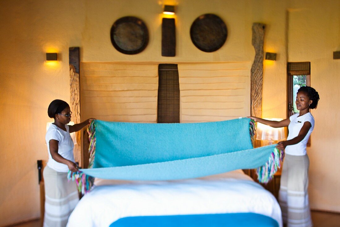Two African women making a bed in front of a yellow wall