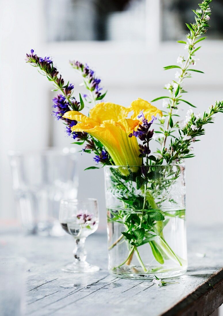 Gelbe Zucchiniblüte und blühende Kräuter in Wasserglas auf altem Tisch