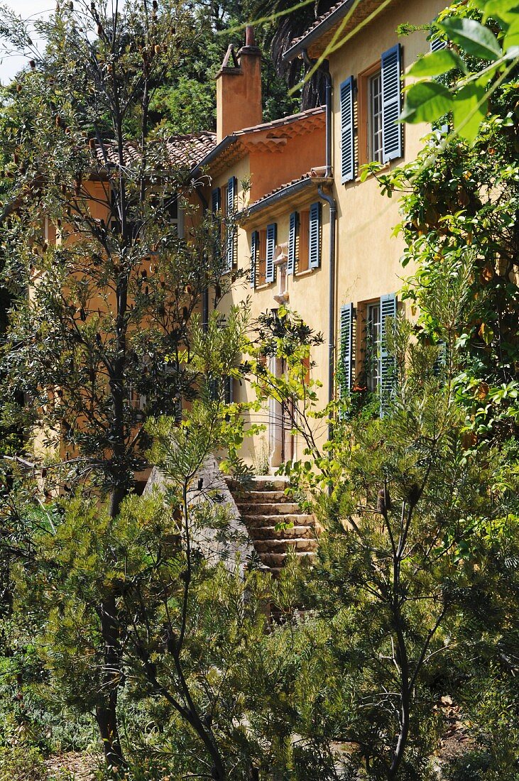 Gelbes Landhaus mit grauen Fensterläden und Terrakottaziegeldach hinter Bäumen versteckt