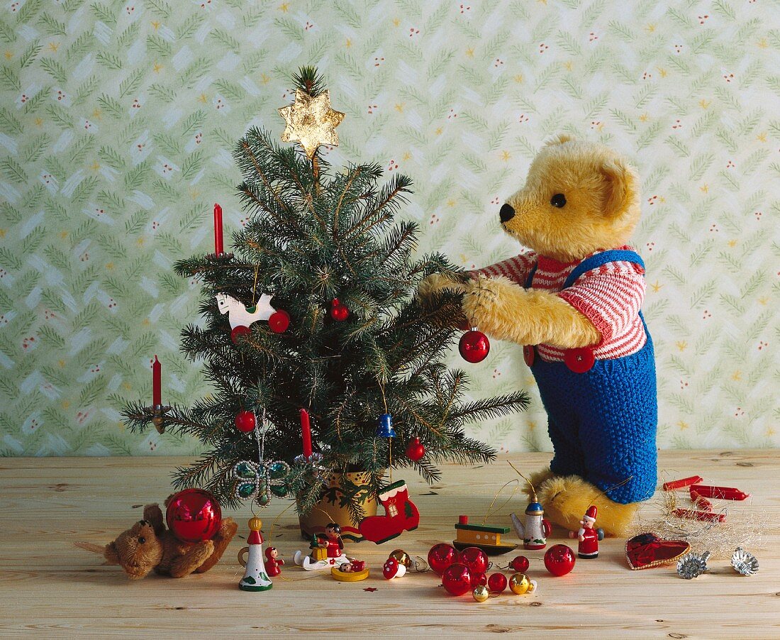 Festive teddy bear decorating little Christmas tree with various baubles lying on floor