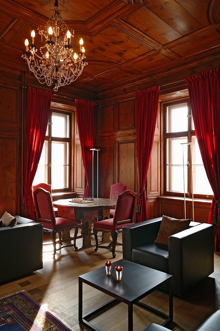 Historischer, holzgetäfelter Salon mit modernen Loungemöbeln und antiker Sitzgruppe vor roten Vorhängen im Hintergrund