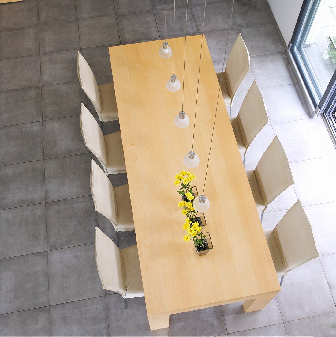 Blick von oben auf Esstisch aus Holz und Stühlen mit hellem Bezug auf hellgrauem Steinboden in minimalistischem Ambiente