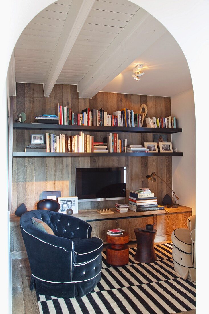 View through arched doorway into room with white, wood-beamed ceiling and brown, wood-clad wall; charcoal velvet armchair on brown and white striped rug in foreground