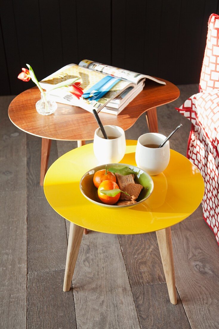 Stacked books, mugs of tea, vase of flowers and fruit bowl on two retro coffee tables