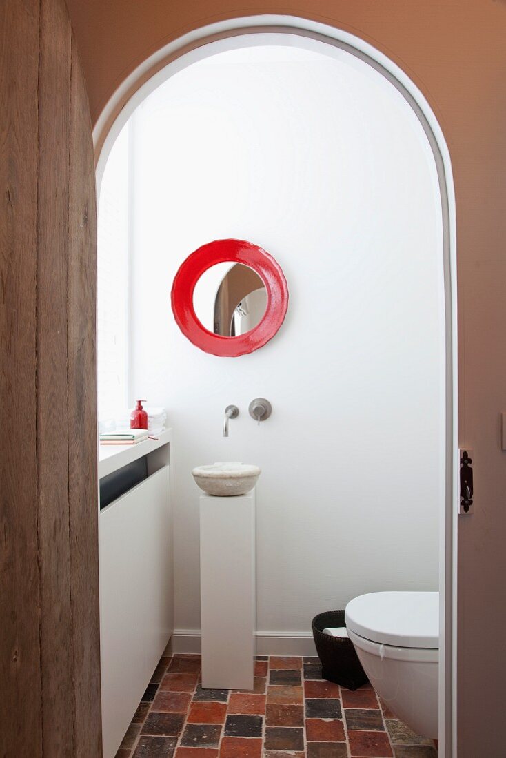 White guest toilet with small pedestal sink below round mirror with red frame and terracotta tiles