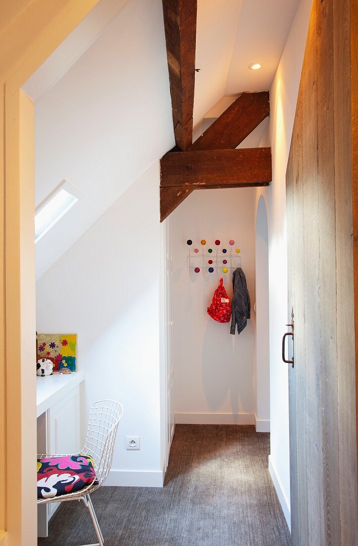 Helles Kinderzimmer mit rustikalen Dachbalken und Schreibtisch unter dem Dachfenster; an der hinteren Wand eine bunte Kindergarderobe
