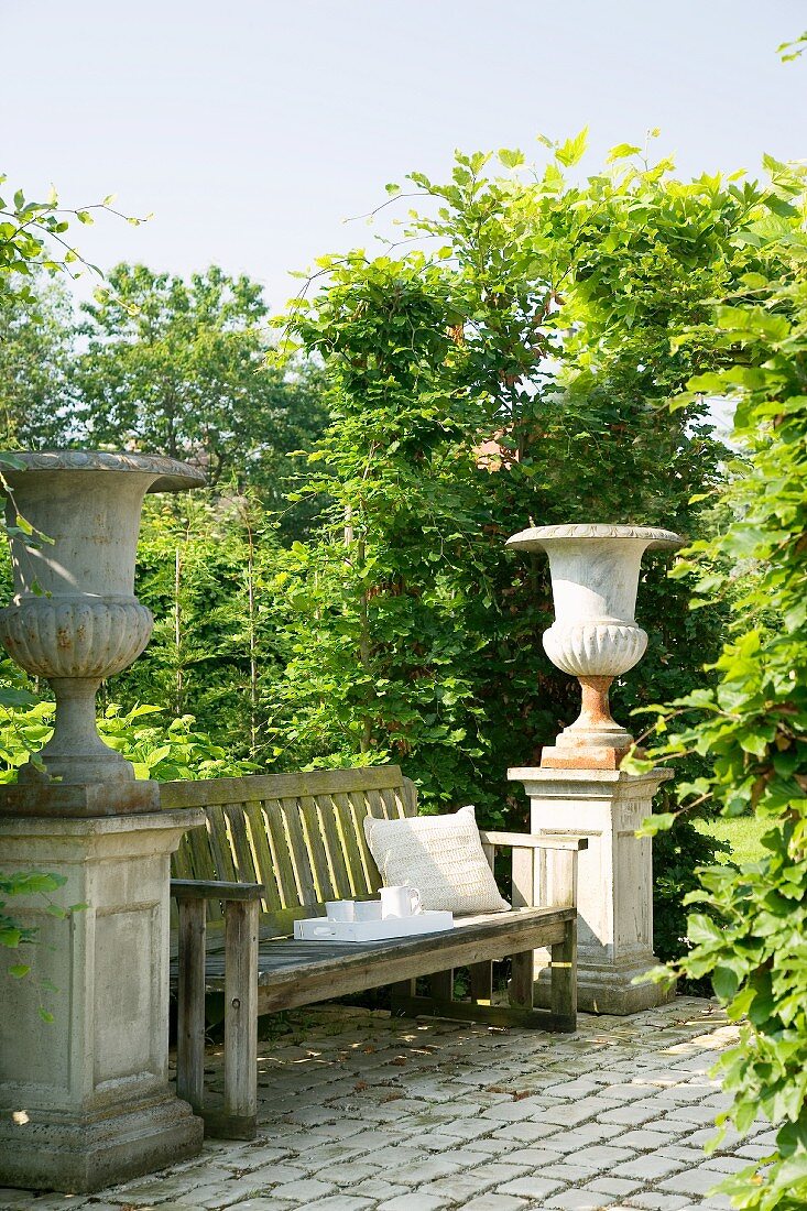 Simple bench flanked by impressive stone urns in sunny garden