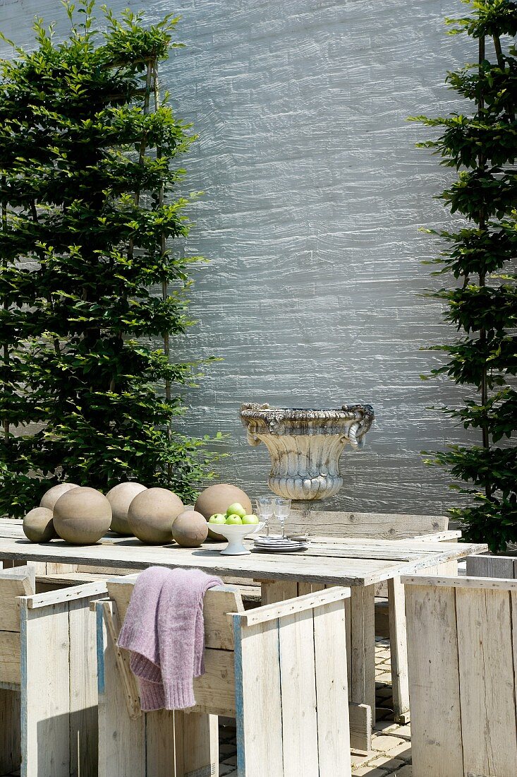 weiße Ziegelsteinwand mit an einem Spalier wachsenden Koniferen; davor ein rustikaler Holztisch mit Stühlen und ein steinerner Pokal
