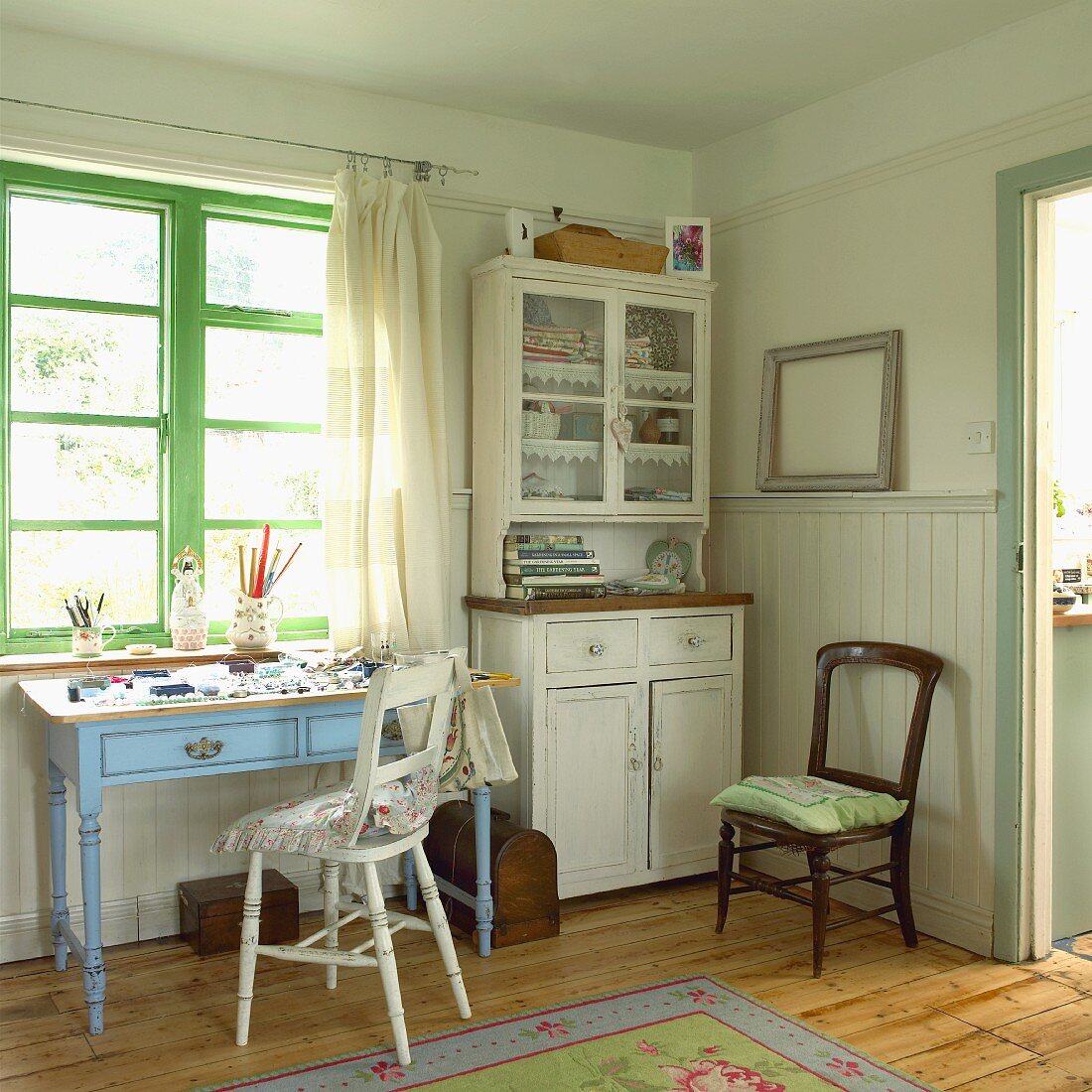 Interior in pastel shades with half-height wooden panelling, glass-fronted dresser and small table with drawers below window with green-painted frame