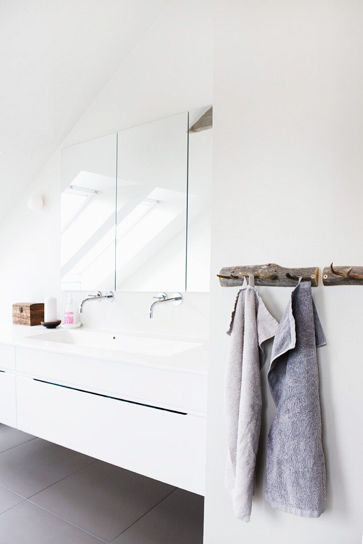 Towels hanging on rustic rack with hooks next to white washstand with wall-mounted fittings below mirrored cabinet