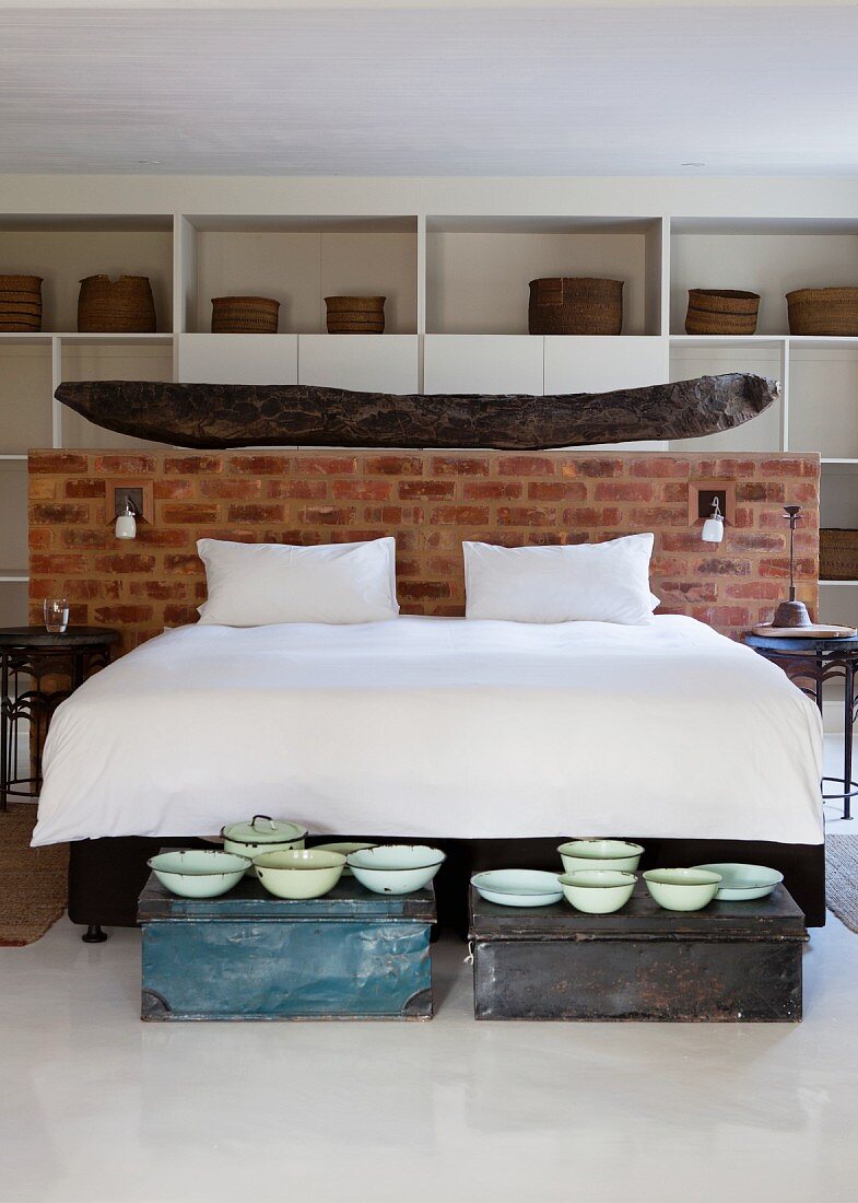 Doppelbett mit weisser Bettwäsche vor halbhoher Ziegelwand und Vintage Schüsseln auf Truhen am Bettende im Schlafzimmer